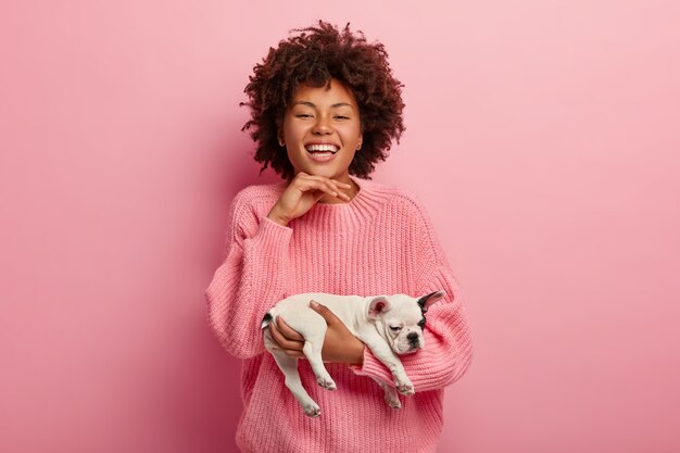 Mujer de aspecto agradable con una gran sonrisa, sostiene la barbilla, lleva un cachorro de pedigrí soñoliento