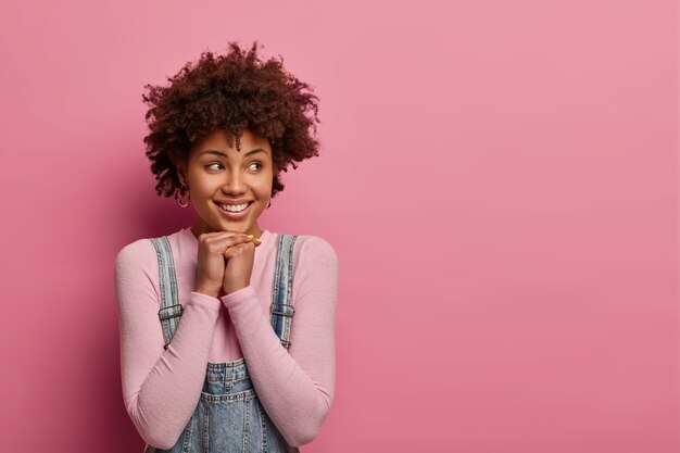 Una mujer de aspecto agradable mantiene las manos debajo de la barbilla, mira hacia otro lado, tiene una sonrisa tierna, viste ropa informal, piensa en algo bueno, posa contra una pared rosa pastel, espacio en blanco para su texto promocional