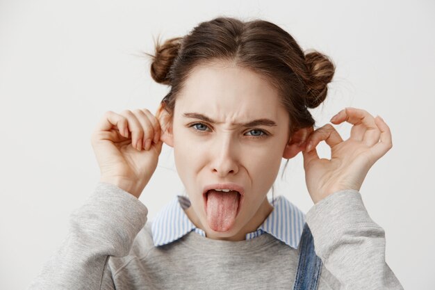 Mujer de aspecto gracioso con una mueca informal con la lengua fuera haciendo que las orejas sobresalgan. Ciérrese encima del retrato del alborotador femenino con el peinado de moda que está loco engañando alrededor Concepto de alegría