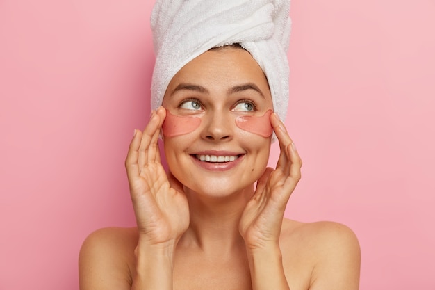 Una mujer de aspecto positivo y agradable se preocupa por la belleza de la piel de los ojos, aplica parches de colágeno humectantes en la cara, mira a un lado, se para en topless, se somete a tratamientos de belleza después del baño, elimina las arrugas