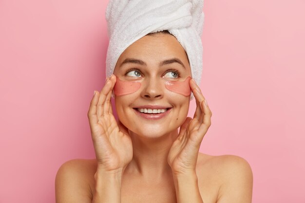 Una mujer de aspecto positivo y agradable se preocupa por la belleza de la piel de los ojos, aplica parches de colágeno humectantes en la cara, mira a un lado, se para en topless, se somete a tratamientos de belleza después del baño, elimina las arrugas