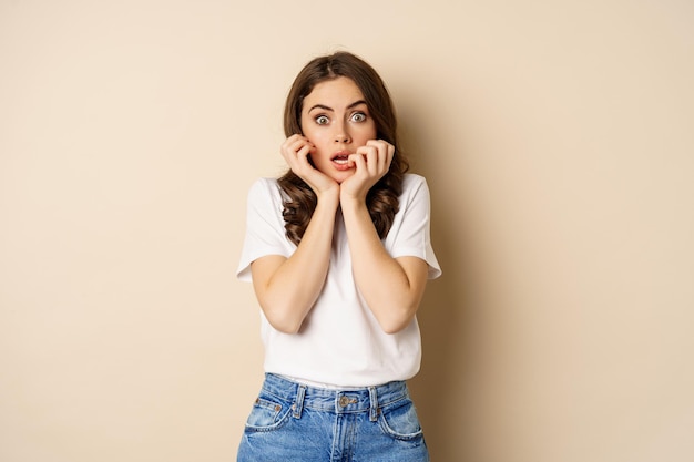 Mujer asustada y conmocionada jadeando, luciendo sorprendida sin palabras, usando una camiseta, de pie sobre un fondo beige.