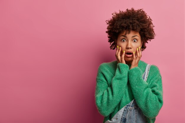 Mujer asustada emocionalmente en pánico, agarra la cara y mira con ojos saltones, se pone de pie alarmada y asustada, mantiene la mandíbula caída, teme a las dificultades, usa un suéter verde, modela sobre una pared rosada