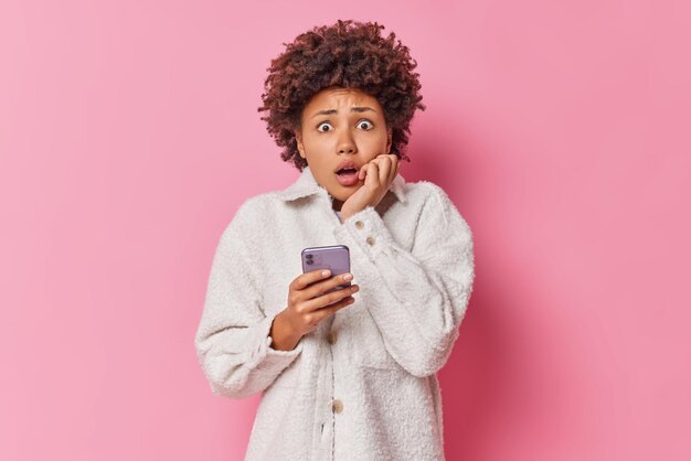 Mujer asustada sostiene celular moderno ve fotos de malas noticias o mensaje tiene expresión temerosa usa poses de abrigo de piel contra la pared rosa