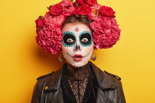 Mujer asustadiza usa maquillaje de Halloween de terror, tiene expresión asustada, chas círculos oscuros pintados alrededor de los ojos, usa una gran corona de flores rojas, aislada sobre fondo amarillo.