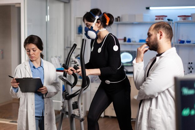 Mujer atleta con máscara en gimnasio bycle trainning resistencia corporal mientras médico investigador mide la frecuencia cardíaca monitoreando los datos de EGK en el laboratorio. Deportista con electrodos médicos