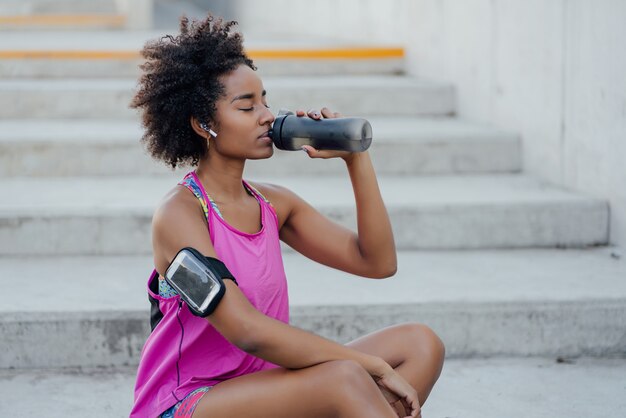 Mujer atlética afro bebiendo agua y relajarse después de hacer ejercicio mientras está sentado en las escaleras al aire libre