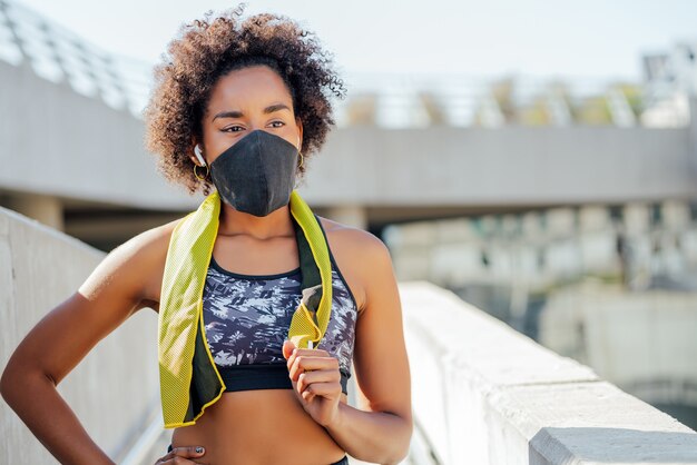 Mujer atlética afro con máscara facial y relajarse después de hacer ejercicio al aire libre en la calle