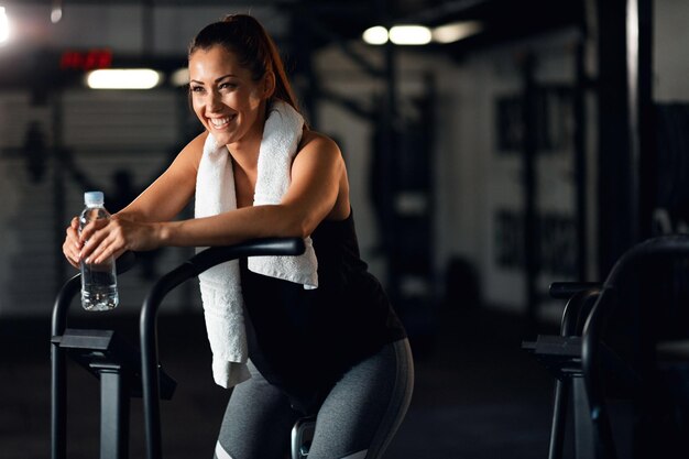 Mujer atlética feliz con una botella de agua tomando un descanso y descansando en una bicicleta estática después de andar en bicicleta en un gimnasio Copiar espacio