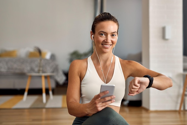 Mujer atlética feliz comprobando la hora en el reloj de pulsera mientras usa un teléfono inteligente y mira la cámara