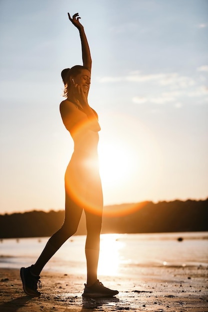 Mujer atlética feliz escuchando música con auriculares mientras hace ejercicio en la orilla del río al amanecer.