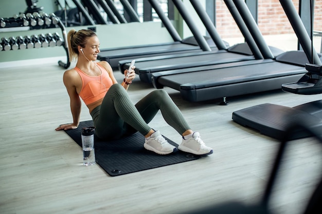 Mujer atlética feliz escuchando música en auriculares mientras usa un teléfono inteligente en un gimnasio