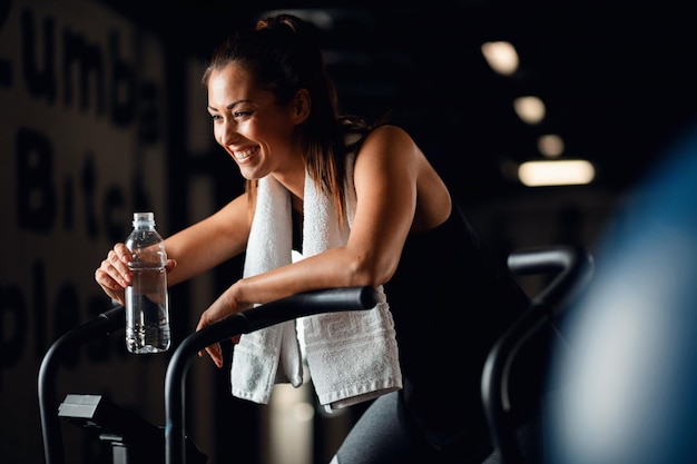Mujer atlética feliz haciendo ejercicio en bicicleta estacionaria y bebiendo agua en un gimnasio