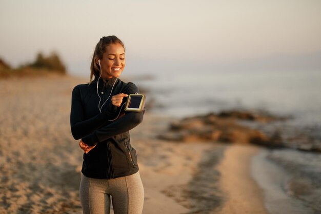 Mujer atlética feliz preparándose para la carrera y ajustando la música en su teléfono inteligente en la playa