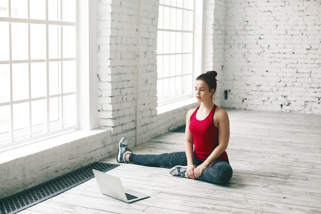 Mujer atlética joven con estilo en leggings, zapatillas y zapatillas para correr sentado en el piso en casa frente a una computadora portátil genérica abierta mientras ve el entrenamiento de yoga en línea, hace varias asanas, tiene una mirada seria