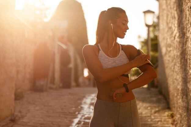 Mujer atlética joven que ajusta la música en su teléfono inteligente y se prepara para el entrenamiento diario en la ciudad