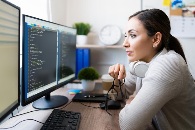 Mujer atractiva de unos 20 años sentada frente a su escritorio y quitándose las gafas mientras lee el código del software