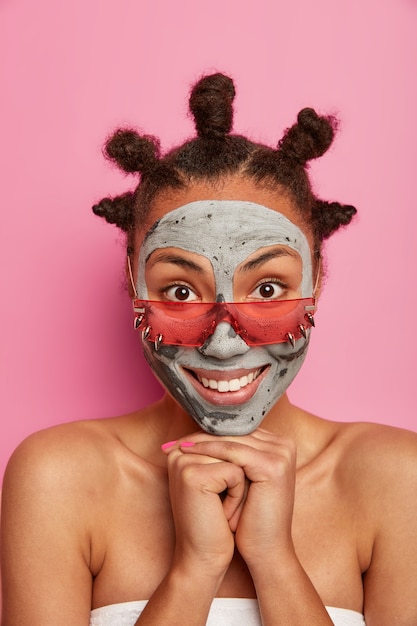 Mujer atractiva aplica mascarilla de barro en la cara, sonríe suavemente, tiene dientes blancos perfectos, piel bien cuidada, usa lentes de sol color de rosa, muestra los hombros desnudos, envuelta en una toalla de baño, se somete a procedimientos de belleza