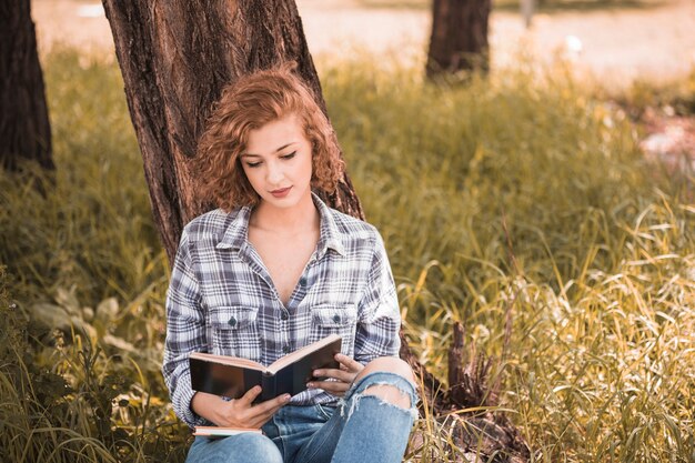 Mujer atractiva apoyándose en el árbol y libro de lectura