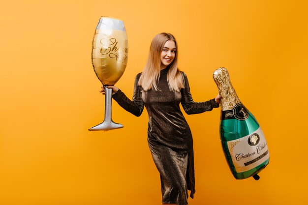 Mujer atractiva bailando en naranja con botella de champagne. Retrato interior de jocund mujer caucásica celebrando un cumpleaños con vino.