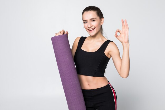 Mujer atractiva con bien en atuendo de gimnasio sosteniendo una estera en sus brazos extendidos aislado en la pared blanca