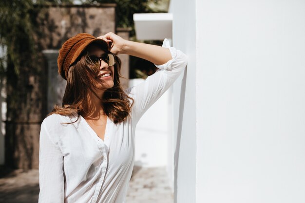 Mujer atractiva con cabello rubio oscuro con gorra y blusa ligera se inclinó sobre la pared del edificio blanco.