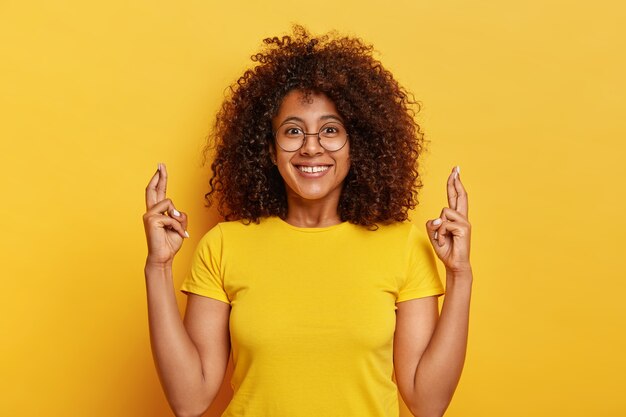 Mujer atractiva en camiseta amarilla, cruza los dedos, espera en un futuro afortunado, sonríe agradablemente, posa sobre un fondo vivo, reza interior