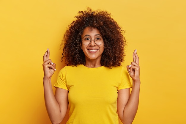 Mujer atractiva en camiseta amarilla, cruza los dedos, espera en un futuro afortunado, sonríe agradablemente, posa sobre un fondo vivo, reza interior