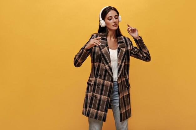 Mujer atractiva en chaqueta elegante y jeans escucha música en auriculares Chica bonita en poses de camiseta blanca sobre fondo amarillo aislado