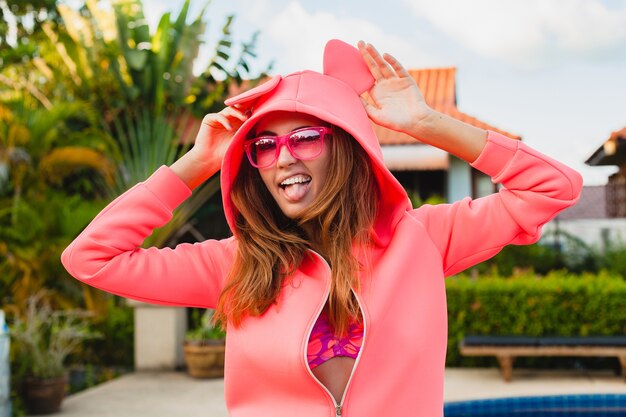 Mujer atractiva en colorida sudadera con capucha rosa con gafas de sol en vacaciones de verano sonriendo expresión de la cara emocional divirtiéndose, estilo de moda deportiva