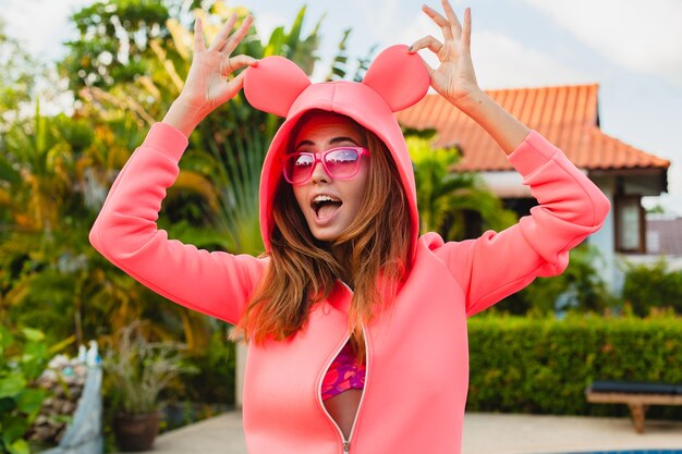 Mujer atractiva en colorida sudadera con capucha rosa con gafas de sol en vacaciones de verano sonriendo expresión de la cara emocional divirtiéndose, estilo de moda deportiva