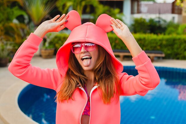 Mujer atractiva en colorida sudadera con capucha rosa con gafas de sol en vacaciones de verano sonriendo expresión de la cara emocional divirtiéndose, estilo de moda deportiva