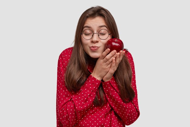 Mujer atractiva complacida con cabello oscuro, mantiene la manzana roja cerca de la mejilla, cierra los ojos, tiene placer, usa anteojos redondos