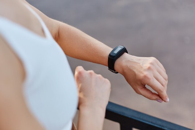 Mujer atractiva delgada sin rostro con top deportivo blanco que controla el dispositivo portátil de seguimiento de la salud y el estado físico, hace ejercicio, entrenamiento al aire libre, estilo de vida saludable