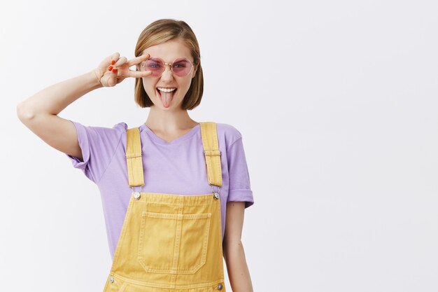 Mujer atractiva descarada con elegantes gafas de sol y ropa de verano que muestra la lengua y el gesto de paz