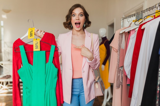 Mujer atractiva emocional sosteniendo coloridos vestidos en percha en tienda de ropa