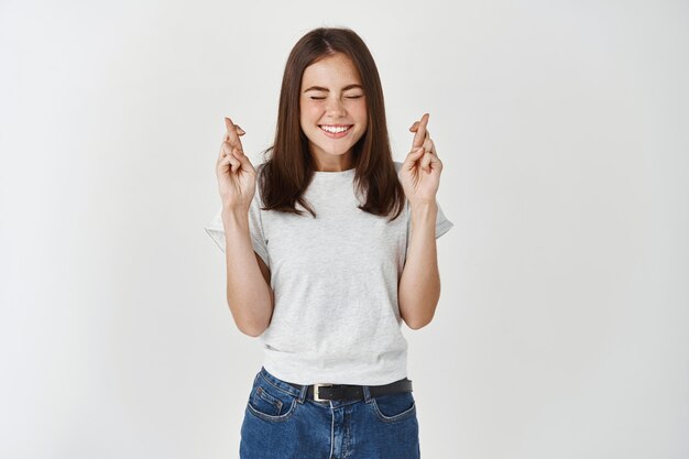 Mujer atractiva entusiasta y emocionada en camiseta, mantenga los dedos cruzados y sonriendo con anticipación en los ojos, los sueños se hacen realidad, pide deseos, anticipa resultados importantes esperanzados