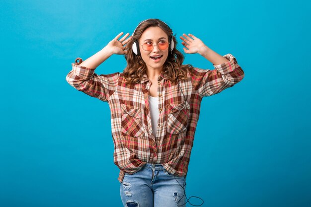 Mujer atractiva en estado de ánimo alegre y expresión de la cara feliz escuchando música en auriculares con camisa a cuadros y jeans aislados sobre fondo azul de estudio