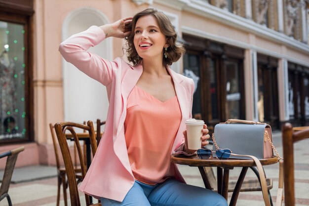 Mujer atractiva en estado de ánimo romántico sonriendo de felicidad sentada en la mesa con chaqueta rosa, ropa elegante, esperando novio en una cita en el café, bebiendo capuchino, expresión de la cara salida