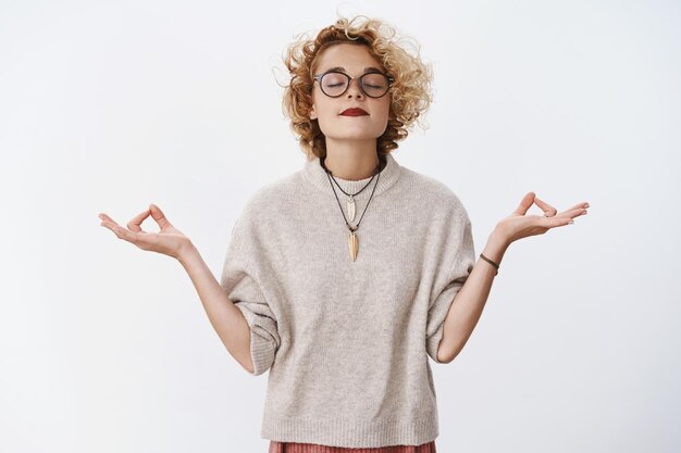 Mujer atractiva con estilo hipster en gafas con ojos cerrados de pelo rubio corto y rizado levantar la cabeza y sonreír como si estuviera de pie en posición de loto con orbes de yoga.