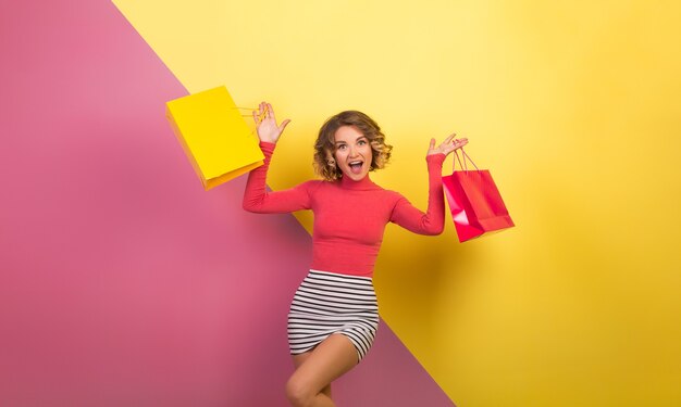 Mujer atractiva excitada en traje elegante y colorido sosteniendo bolsas de la compra con expresión de cara feliz emocionada, fondo amarillo rosa emocional, cuello polo, minifalda a rayas, venta, discout, adicto a las compras