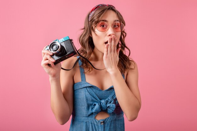 Mujer atractiva con expresión de la cara emocional sorprendida divertida con cámara vintage en vestido de mezclilla y gafas de sol sobre fondo rosa