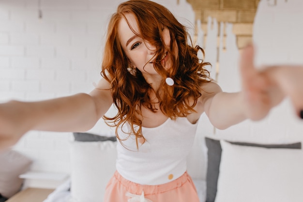 Mujer atractiva feliz haciendo selfie en casa. Magnífica chica de jengibre con peinado romántico posando en su habitación.