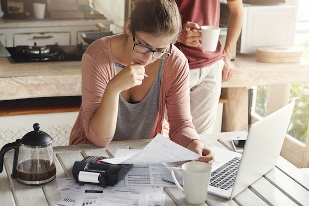 Mujer atractiva con gafas que tiene una mirada seria y concentrada sosteniendo un bolígrafo mientras llena papeles, calcula facturas, reduce los gastos familiares, intenta ahorrar dinero para hacer una gran compra