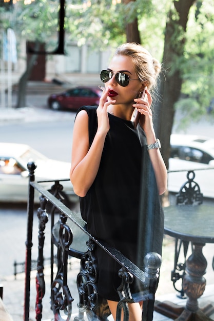 Mujer atractiva en gafas de sol en vestido corto negro fumando cigarrillos en la terraza. Ella está hablando por teléfono, mirando a un lado.