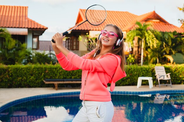 Mujer atractiva haciendo deporte en la piscina en colorida sudadera con capucha rosa con gafas de sol escuchando música en auriculares en las vacaciones de verano, jugar al tenis, estilo deportivo