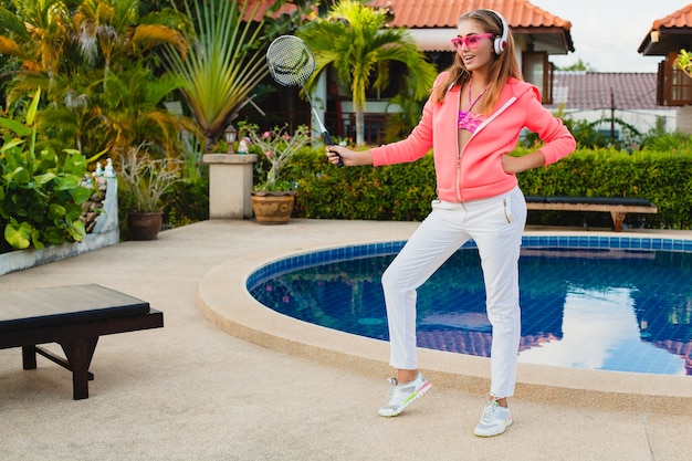 Mujer atractiva haciendo deporte en la piscina en colorida sudadera con capucha rosa con gafas de sol escuchando música en auriculares en las vacaciones de verano, jugar al tenis, estilo deportivo