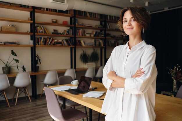 Mujer atractiva joven en camisa blanca mirando soñadoramente en cámara con escritorio en el fondo en la oficina moderna