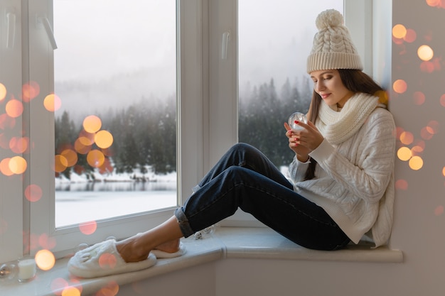 Mujer atractiva joven en elegante suéter de punto blanco, bufanda y sombrero sentado en casa en el alféizar de la ventana en Navidad con decoración de presente de bola de nieve de cristal, vista del bosque de invierno, luces bokeh