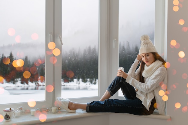 Mujer atractiva joven en elegante suéter de punto blanco, bufanda y sombrero sentado en casa en el alféizar de la ventana en Navidad con decoración de presente de bola de nieve de cristal, vista del bosque de invierno, luces bokeh